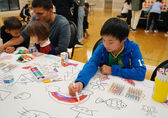 A boy draws a rainbow. At the table next to him, a man draws with two younger children leaning on him and watching him.